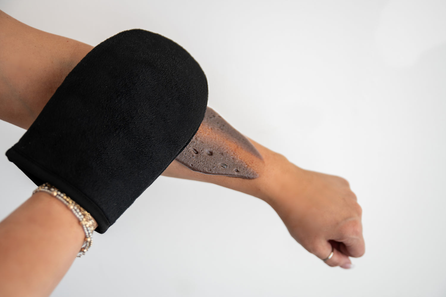 A black application mitt applying a dark tanning mousse to an arm in front of a white background.