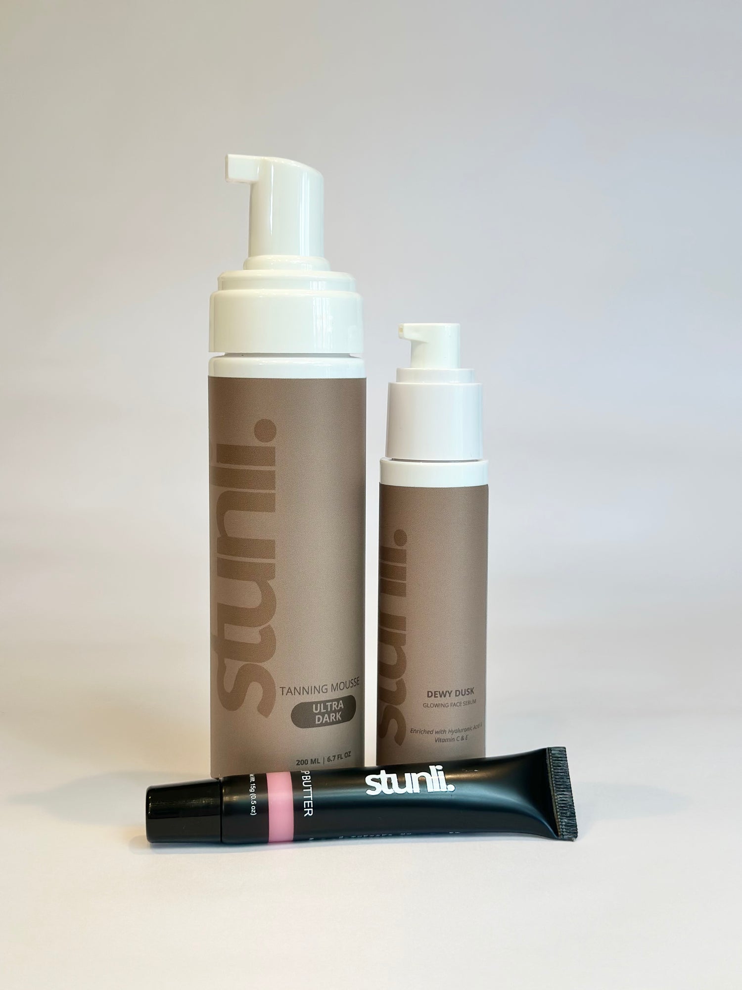 A set of three Stunli brand cosmetic products, including a tanning mousse, a serum, and a lip butter, with packaging in shades of pink, black, and beige.
