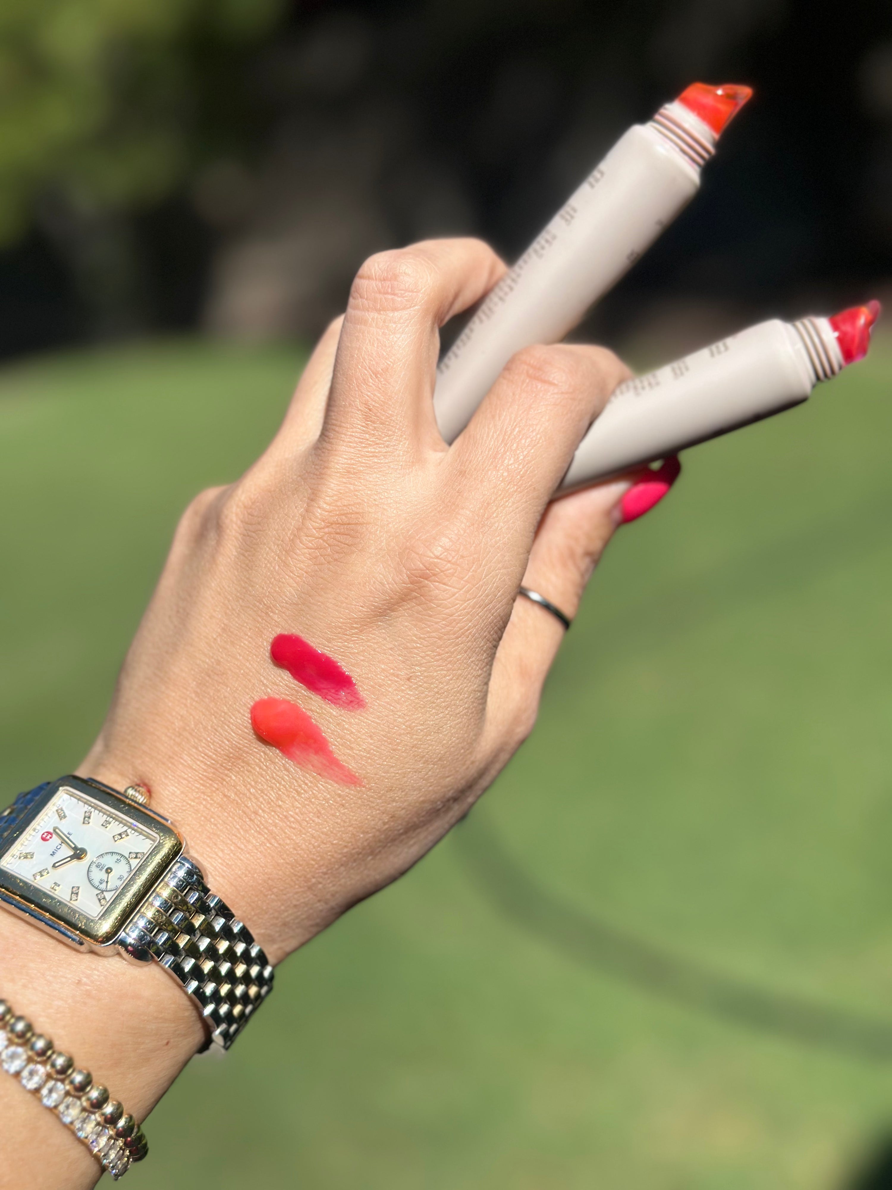 A close-up of a hand holding two cosmetic markers with coral-red tips, showing two lipstick-like color swatches on the back of the hand. The person is wearing a silver watch and bracelet, with a blurred green outdoor background.