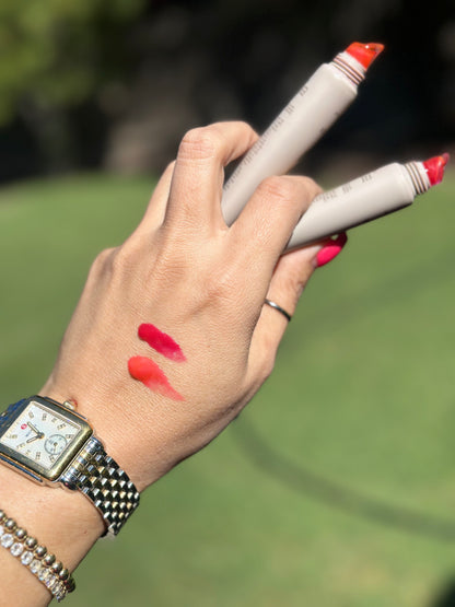 A close-up of a hand holding two cosmetic markers with coral-red tips, showing two lipstick-like color swatches on the back of the hand. The person is wearing a silver watch and bracelet, with a blurred green outdoor background.