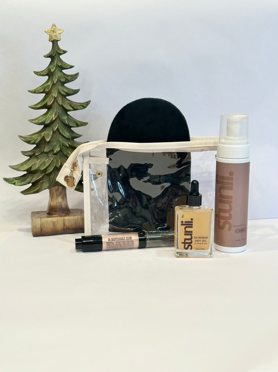 Cosmetic products including a bottle, brush, and jar on a white surface with a small decorative tree in the background.