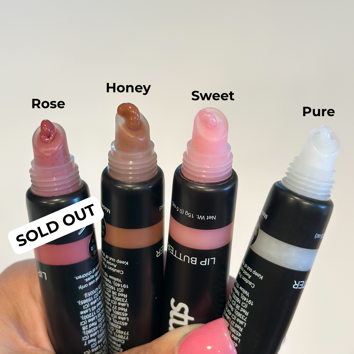 Four lip butter tubes labeled Rose, Honey, Sweet, and Pure are shown in a hand against a plain light background. The tubes are black with different shade caps visible, and a white &